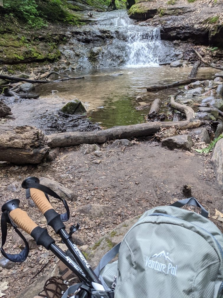 An idyllic, small waterfall flows into a pool of water in the background. In the foreground is a full backpack, hiking poles, and hiking boots. Taken at Palfrey's Glen.